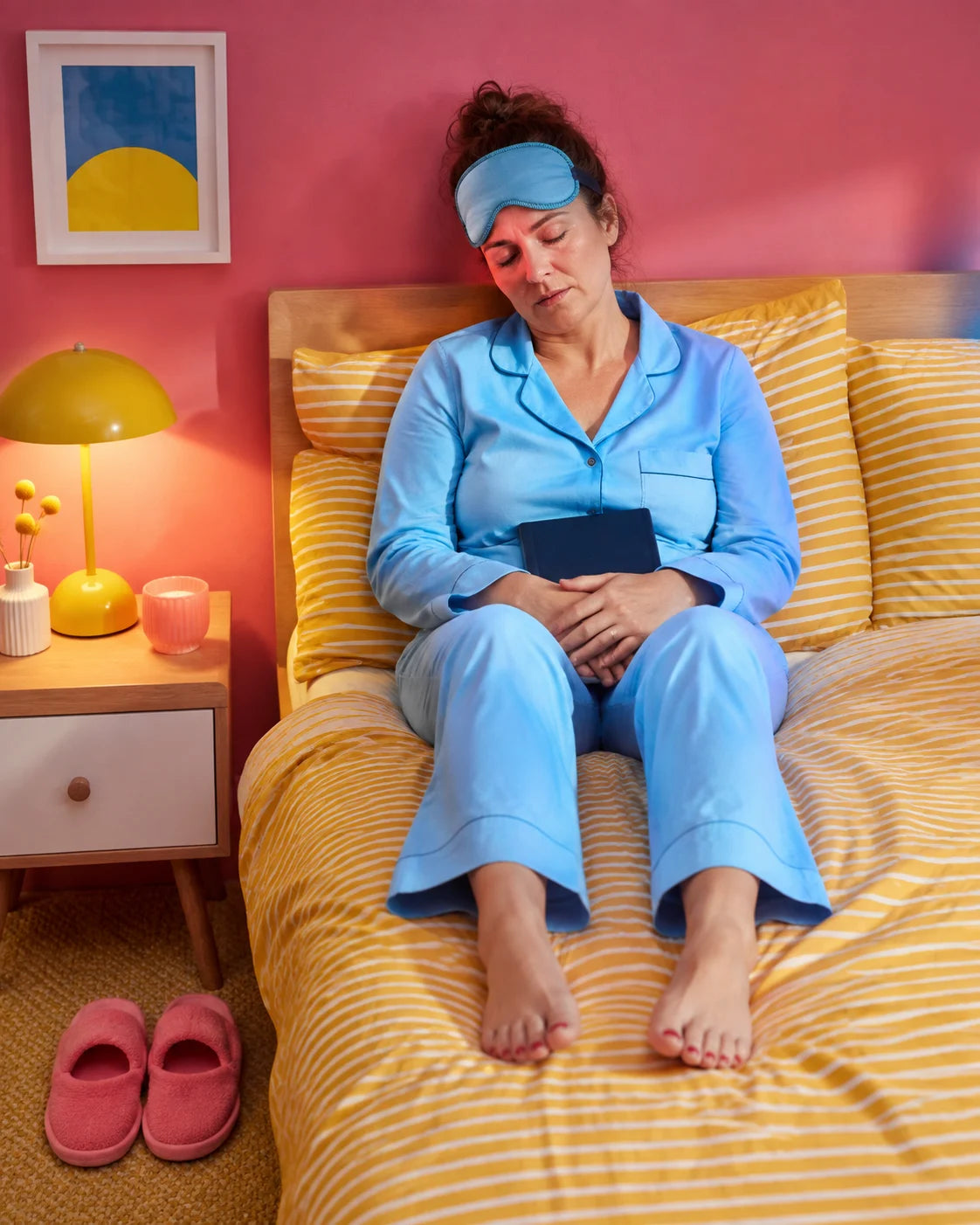 A woman in blue pajamas has dozed off with a closed book on her lap.