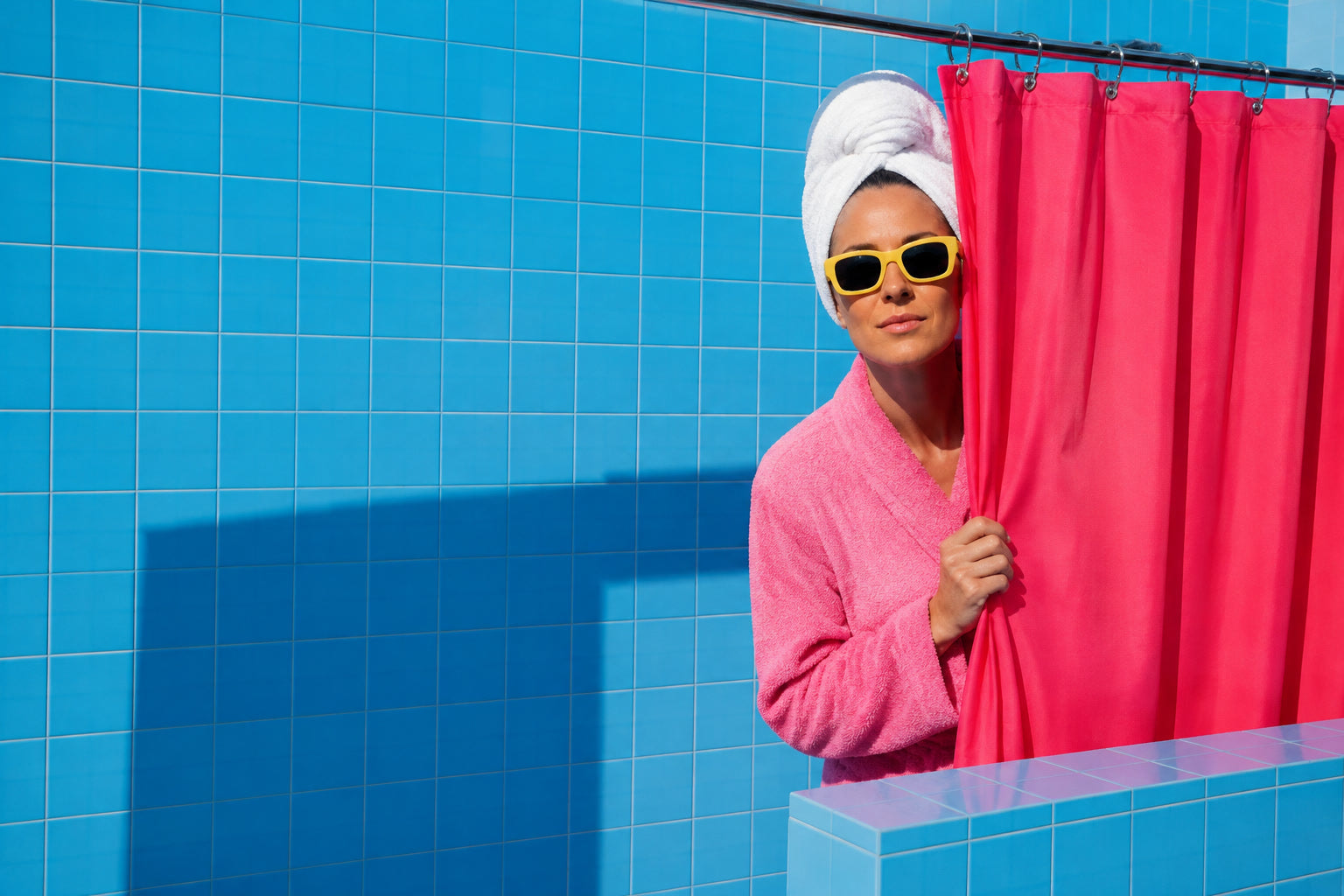 An adult in a towel turban and yellow sunglasses peeks around a bright pink shower curtain.