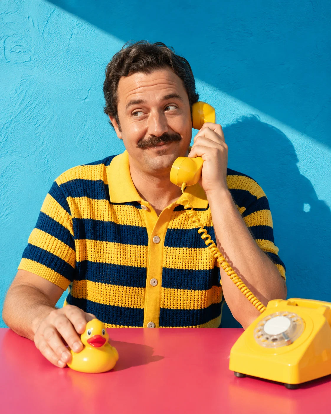 A man takes a call on a yellow rotary phone, with a rubber duck as his desk assistant.