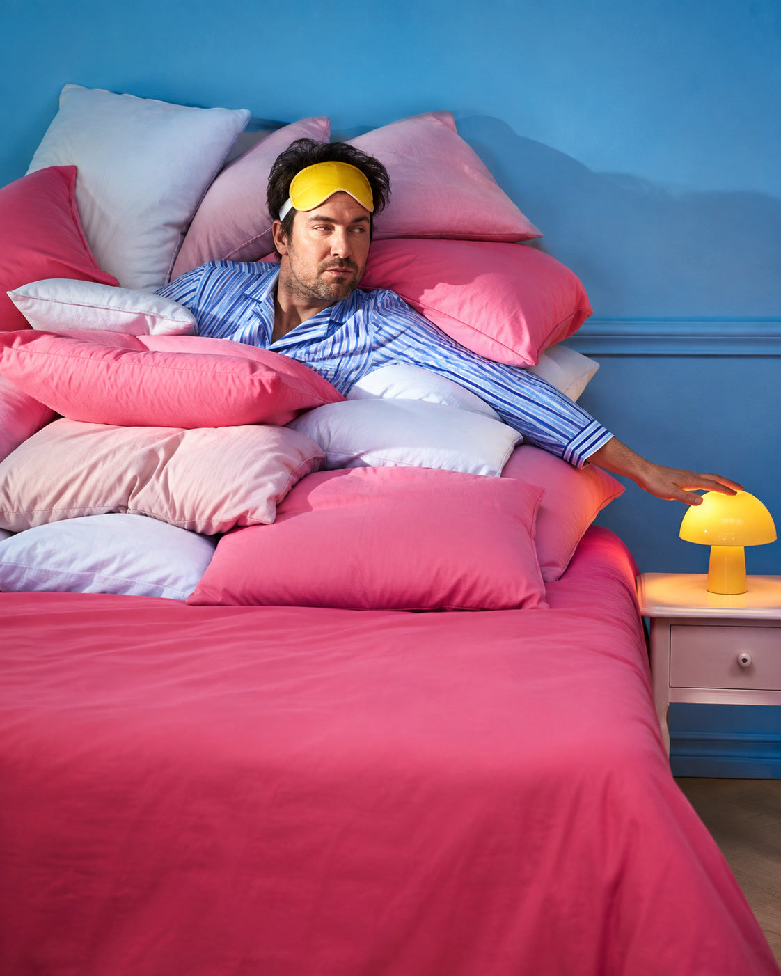 An adult in striped pajamas reaches for a lamp from an enormous pile of pink pillows.